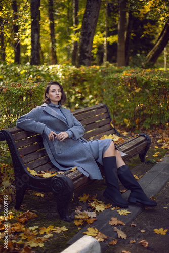 brunette woman in a gray coat and boots is sitting on a park bench in autumn among the trees. Orange foliage and a wooden bench during the day