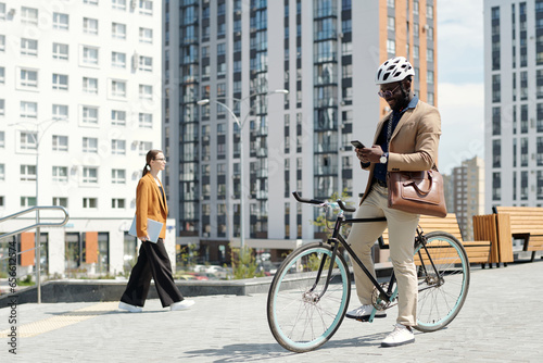 Young smiling male entrepreneur in safety helmet sitting on bicycle and scrolling through online information in mobile phone in the street photo