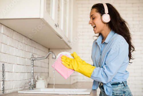 Fotografie Joyful woman wearing wireless headphones and singing while washing dishes in kit