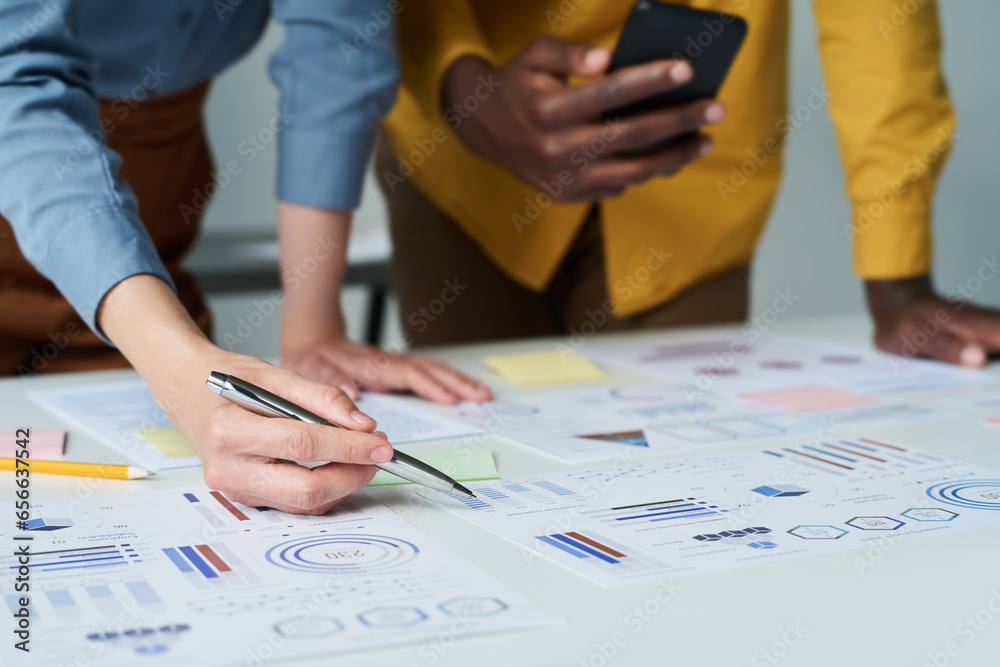 Focus on hand of female economist or accountant pointing at financial document while discussing chart with male colleague with smartphone Focus on hand of female economist or accountant pointing at financial document while discussing chart with male colleague with smartphone