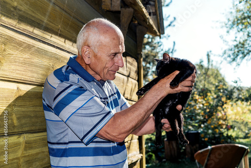Elderly man holds pet black kitty in hands at home yard against yellow wooden wall outdoor