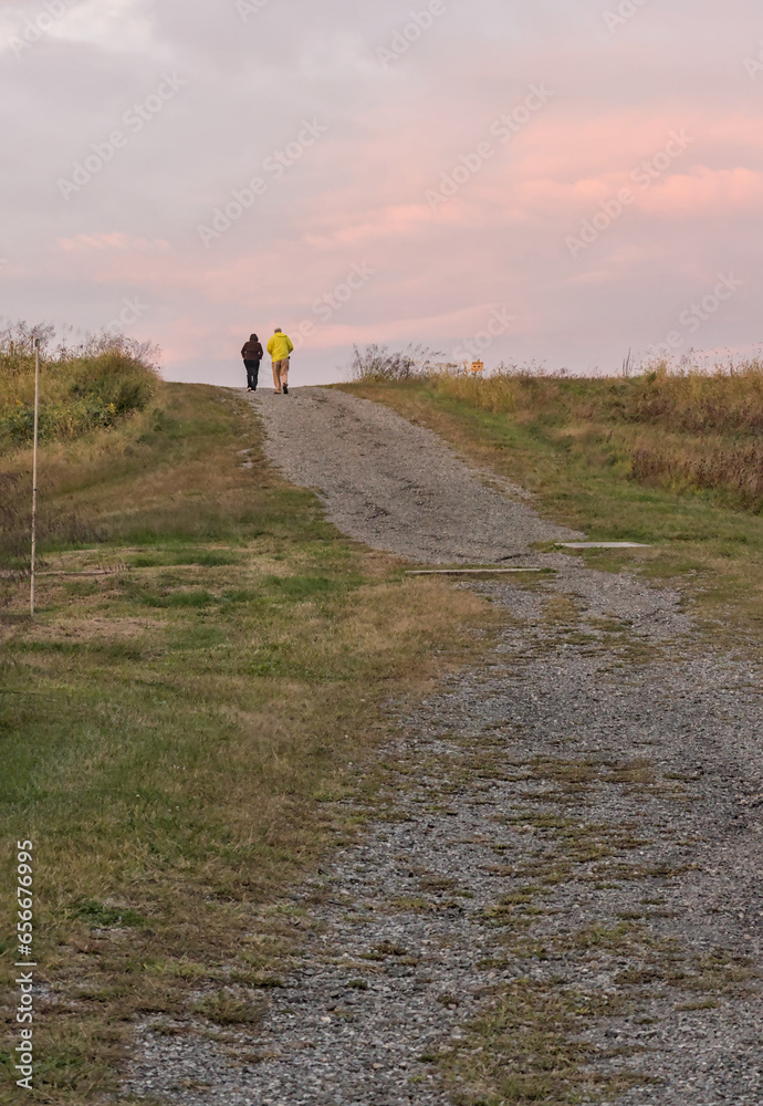 older couple walking on a gravel path on a hill at dusk in a state park (elderly, people, not recognizable, nature, hiking, walk, recreation) photo from behind, in the distance