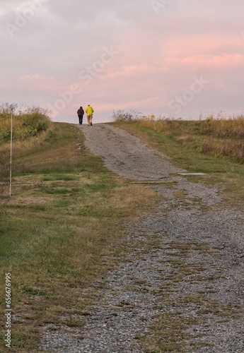 older couple walking on a gravel path on a hill at dusk in a state park (elderly, people, not recognizable, nature, hiking, walk, recreation) photo from behind, in the distance © Yuriy T
