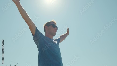 Happy relaxed man in sunglasses breathing fresh air raising arms over blue sky with sun. Dreaming, freedom and summer travelin photo