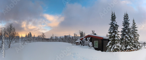 A small snow covered hunting lodge in the norwegian mountains with animal traces in the foreground and mountain summits in the background