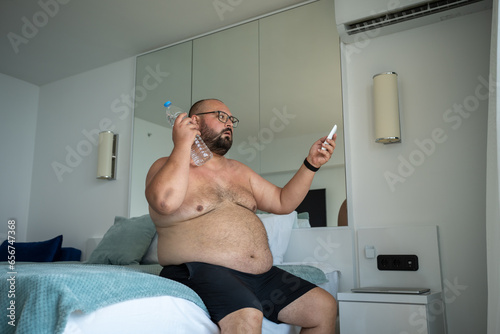 Принти на полотні Naked obese man with bottle of cold water sweats lot adjusts air conditioner with remote control trying to cope with abnormal degrees of warmth sitting in hotel room