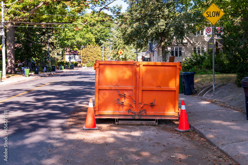 Photography A roll-off construction dumpster parked on a residential street with traffic cones on a shady street in an older neighourhood