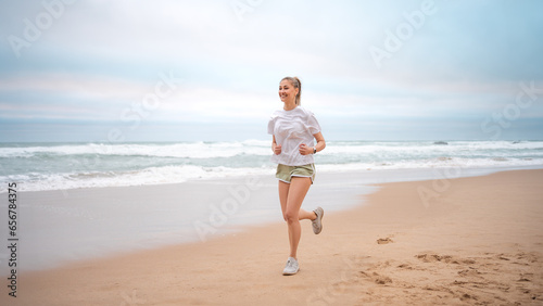 Fototapeta Naklejka Na Ścianę i Meble -  Woman jogging on beach. Sporty female jogging on sand beach, waves on background. Full body view woman in mini shorts and t-shirt who runs alongside ocean. Outdoor workout of trained fit girl. 