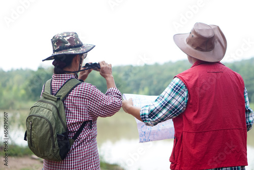 Back view of Asian woman and man travellers holds binocular and paper map to explore natural route for trekking beside the lake. Ecology study. Pastime activity, lifestyle. Explore environment 