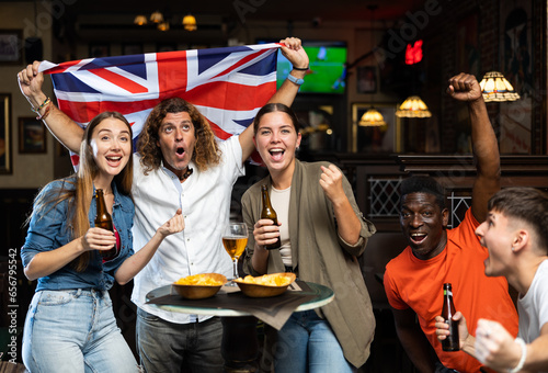 Happy multiracial friends holding the flag of Great Britain, supporting national football team in the bar