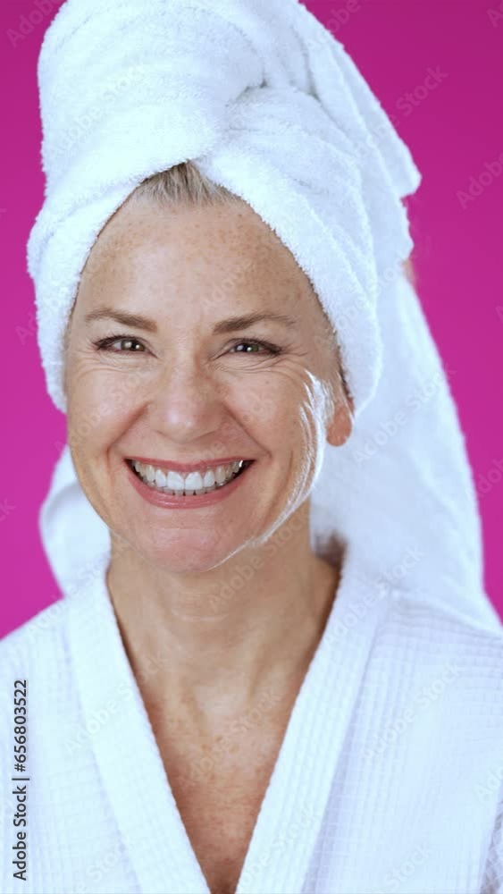Vertical video of attractive mature woman 50s, 60s, wearing bathrobe, looks at camera gives mind blown gesture showing explosion of ideas posing on solid pink background.