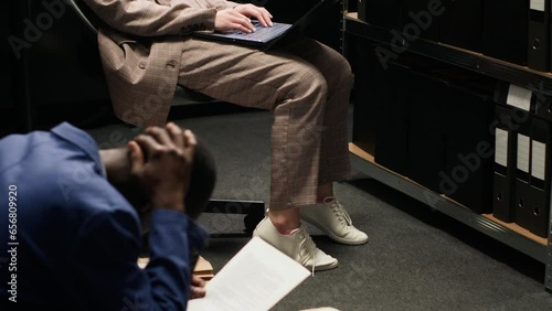 African american male officer and caucasian female inspector with laptop analyze evidence in incident room. Tired policewoman sits on chair while policeman on the floor examining clues and statements. photo