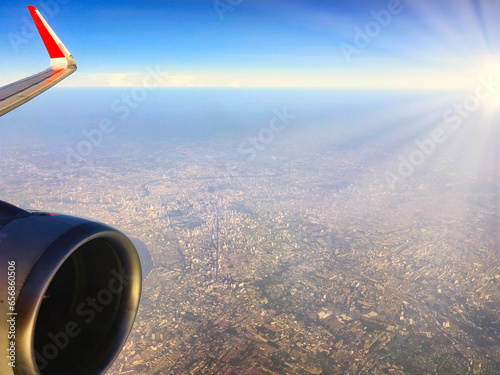 Aerial view of aeroplane jet engine and partial wing over urban city down below with sun ray beam at the horizon