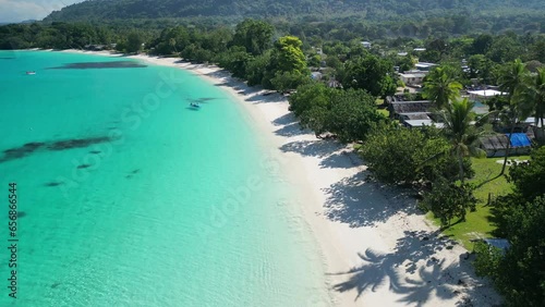 A drone flies over a stunning white sand beach with a red boat at Port Olry on the island of Espiritu Santo. The untouched area in remote Vanuatu is known for its green hills and stunning blue water.