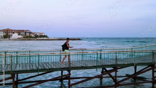 Side view of young man with a backpack and sunglasses walking on a wooden pier at Robinson Beach on the Bulgarian Black Sea coast photo