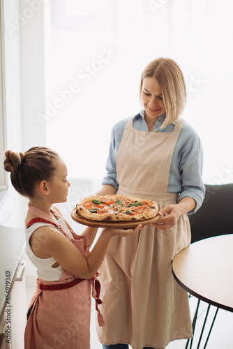 A beautiful mother and her cute daughter are holding a freshly baked pizza. Concept of cooking at home,