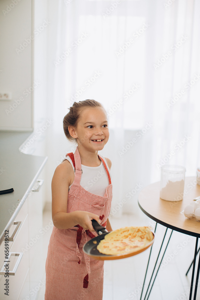 A little 8-year-old girl in an apron is frying pancakes in the kitchen.