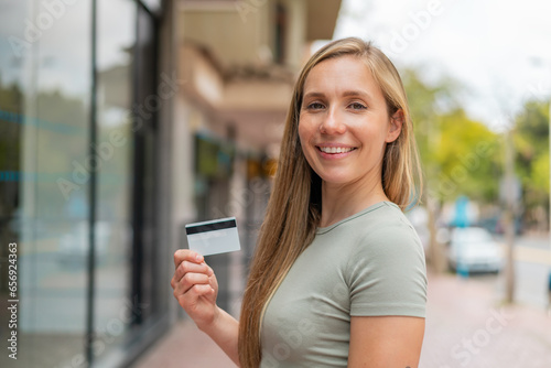 Young blonde woman holding a credit card at outdoors smiling a lot