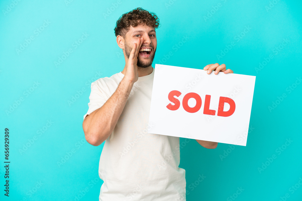 Young handsome caucasian man isolated on blue background holding a placard with text SOLD and shouting Young handsome caucasian man isolated on blue background holding a placard with text SOLD and shouting