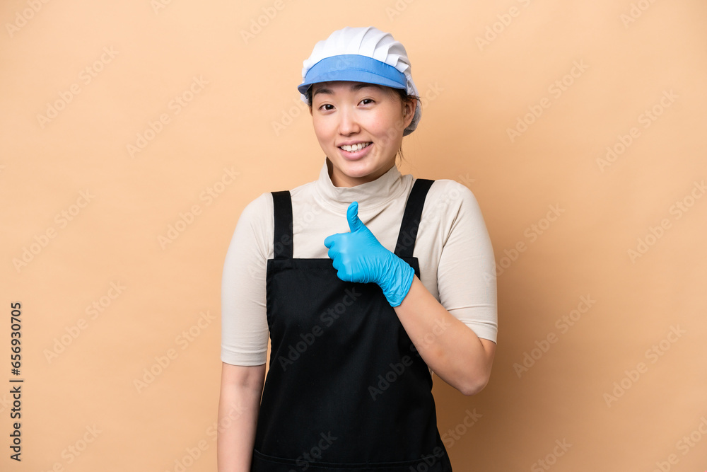 Young Chinese Fishmonger woman wearing an apron and holding a raw fish isolated on pink background giving a thumbs up gesture Young Chinese Fishmonger woman wearing an apron and holding a raw fish isolated on pink background giving a thumbs up gesture