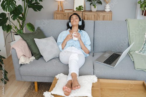 Smiling woman listening to music holding coffee on sofa at home photo