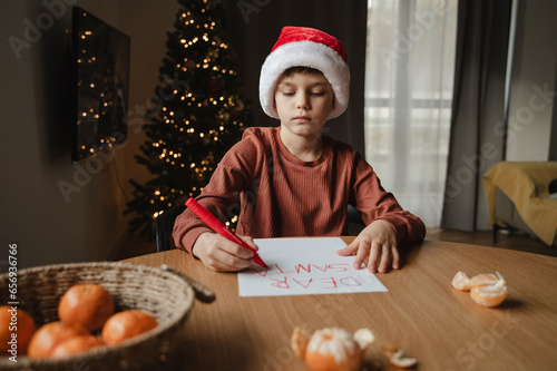 Boy wearing santa hat writing on paper at table photo