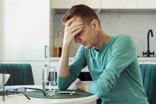 Man is sitting at home at kitchen table and holding his head with his hand. On table is glass of water and pills.