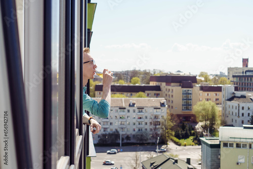 Rest and relaxation. Man with glasses, traveler drinks from glass of juice looks out of apartment window, . City.