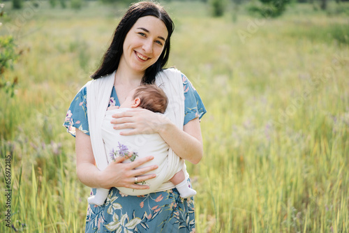 Happy mother holding daughter wrapped in baby sling at meadow photo