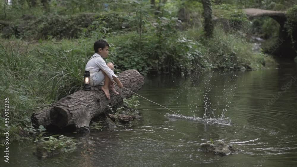 A kid alone n the forest sitting on a tree trunk with a lantern and using a stick to splah water in the river. Wild and free childhood in the woods
