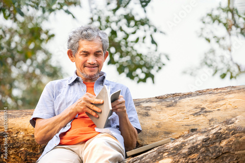 portrait happy senior man sitting and reading a book on tree trunk in summer park,peaceful old mature male relax outside in nature,concept of elderly people lifestyle,hobby,relaxation