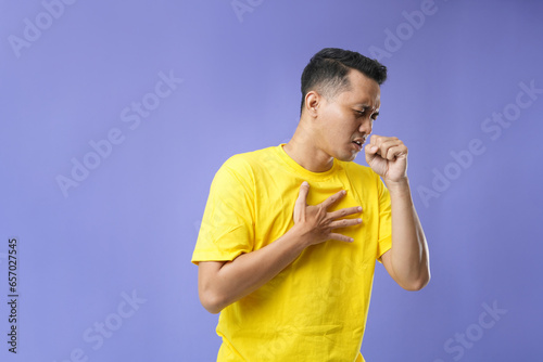 A young Asian man feeling a sore throat and flu symptoms, coughing with his mouth covered. Isolated on a purple background.