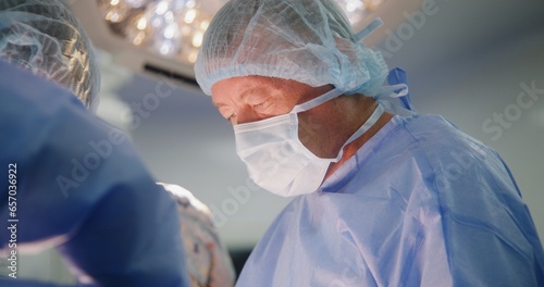 Close up of mature male surgeon in surgical suit operating patient with assistants in surgery room. Professional medics cauterize wound. Modern equipment in operating block. Medical staff at work. photo