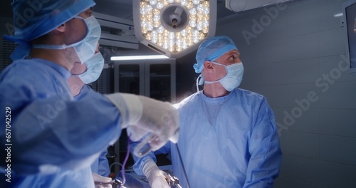 Adult man lies on table under anesthesia. Professional male surgeons operate patient using laparoscopy instruments and looks at monitor. Young nurse assists medics. Medical staff in surgery room.