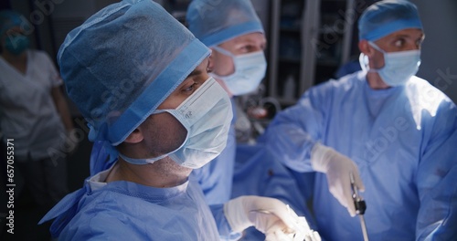 Close up of male surgeon in surgical suit performing laparoscopy operation with colleagues. Professional medics operate patient and look at monitors in modern operating room. Medical staff at work.