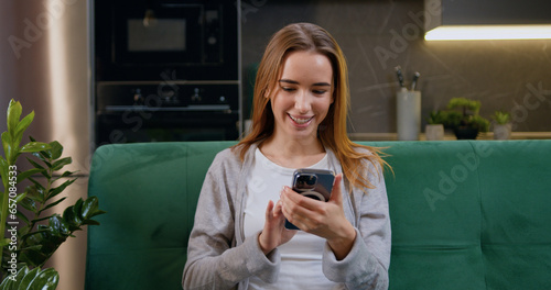 Happy young caucasian woman using mobile phone while sitting a green couch at home kitchen background. Smiling girl with blond hair relaxing on cozy sofa and browsing internet on smartphone