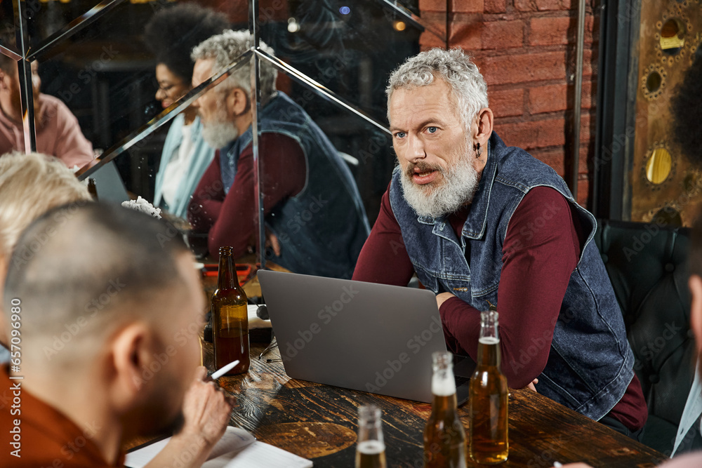 bearded man sitting near laptop and discussing startup project with multiethnic team in bar