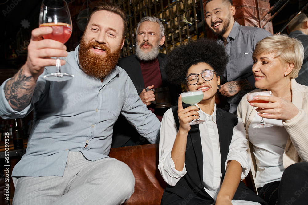 pleased tattooed and bearded man looking at cocktail near multiethnic colleagues with glasses in bar pleased tattooed and bearded man looking at cocktail near multiethnic colleagues with glasses in bar