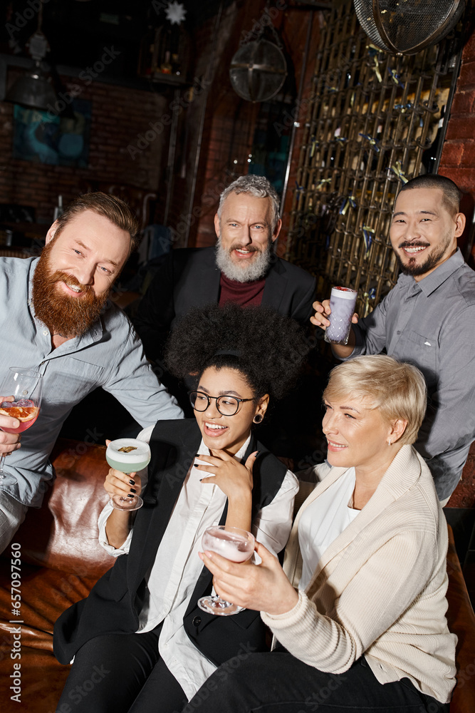 Fototapeta premium overjoyed african american woman laughing near multicultural colleagues with drinks in cocktail bar
