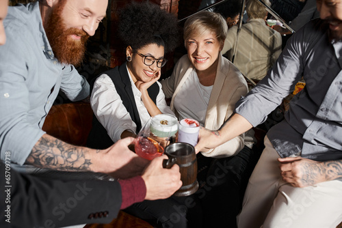cheerful multiethnic colleagues clinking glasses with cocktails while having fun in bar after work photo