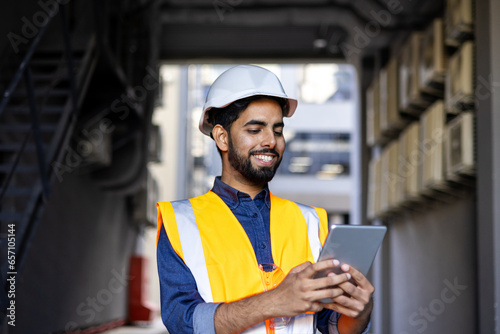 Portrait of young engineer worker, man in helmet and vest using tablet computer, smiling, checking production figures.