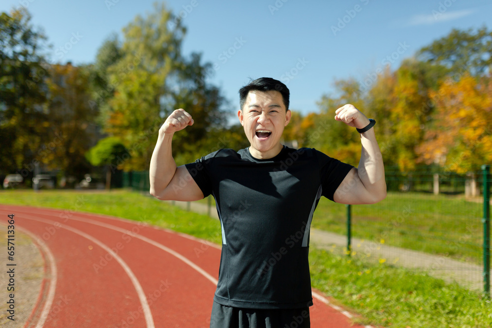 Portrait of a happy young Asian male athlete standing in a stadium, rejoicing at the camera for winning and showing the camera a gesture of strength and victory with his hands.