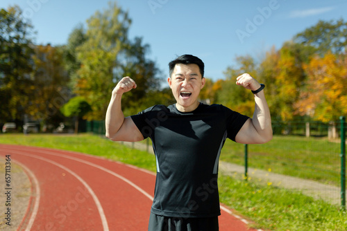 Portrait of a happy young Asian male athlete standing in a stadium, rejoicing at the camera for winning and showing the camera a gesture of strength and victory with his hands.