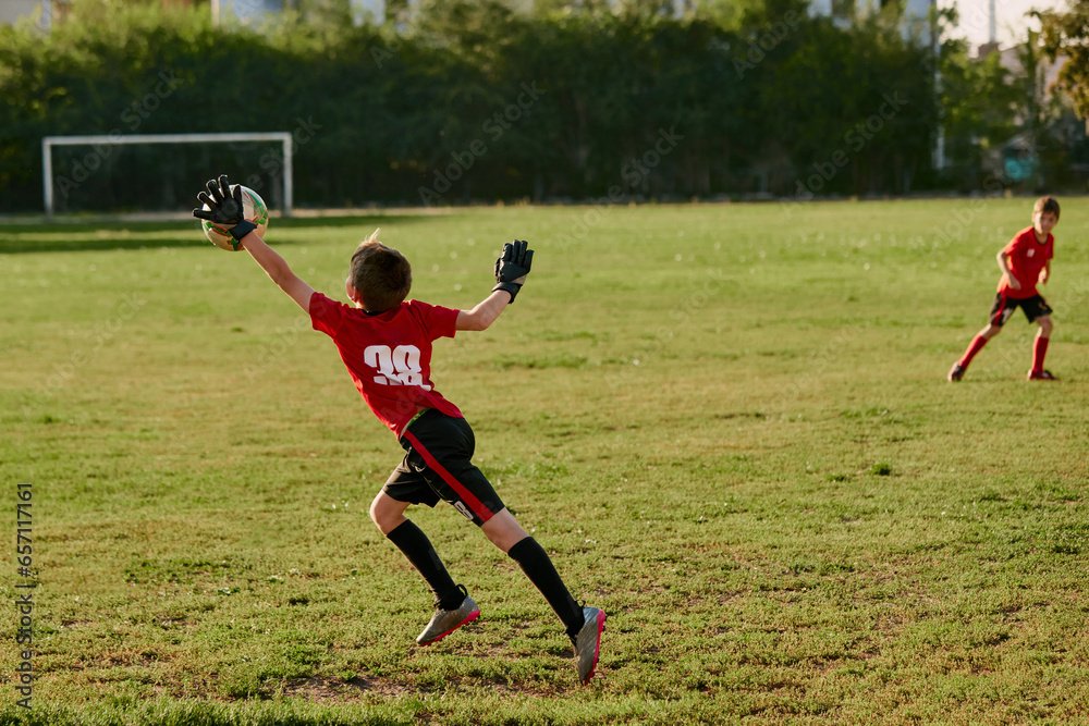 Obraz premium Rear view portrait of goalkeeper, kid beats arm flying ball from kicking boy, striker. Playing football on the sports field. Children's team games.