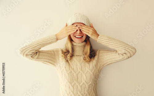 Portrait of cheerful laughing happy young woman covering her eyes with hands in warm soft knitted clothes, sweater and hat on beige background