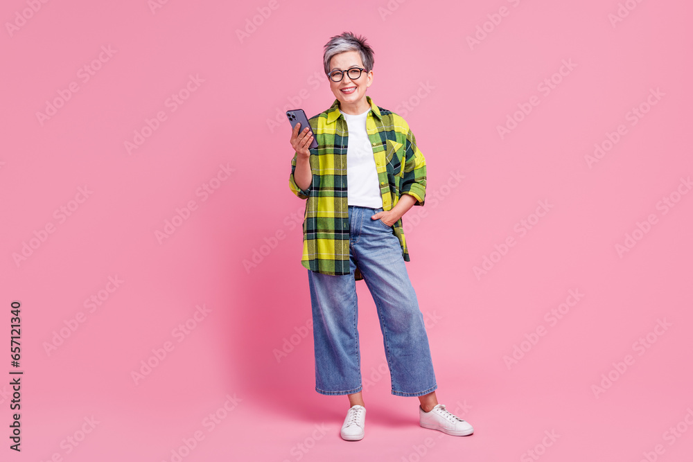 Full length photo of good mood senior woman wear checkered shirt holding smartphone hand in pocket isolated on pink color background Full length photo of good mood senior woman wear checkered shirt holding smartphone hand in pocket isolated on pink color background