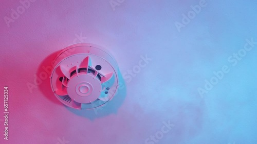 The smoke detector is mounted on the ceiling and the smoke rising underneath is illuminated by flashing red and blue lights.