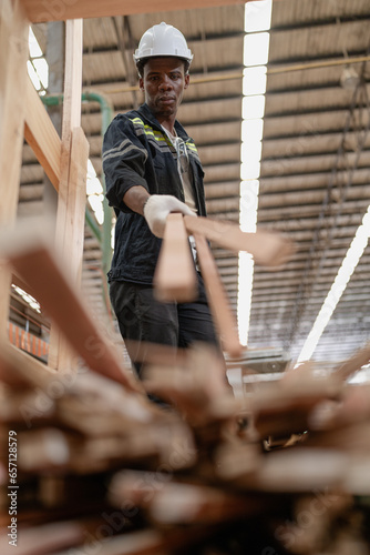 Young adult worker standing in warehouse examining hardwood material for wood furniture production. Male supervisor wears uniform and safety hardhat working in lumber pallet factory. Woodwork industry