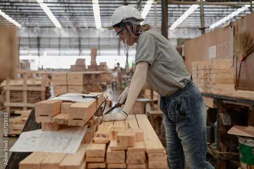 Young adult woman worker working in warehouse examining hardwood material for wooden furniture production. Female technician in safety uniform hardhat working professional job in lumber pallet factory