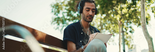 Young italian guy with ponytail and stubble sits with headphones on street bench listens to music and makes sketches with pen on piece of paper on cityscape background